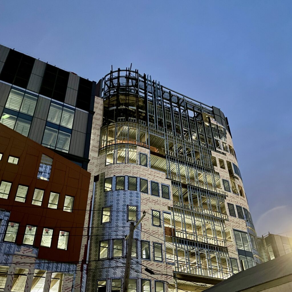 Cold-formed steel (CFS) framing installation progresses along the exterior wall, including hoist infill framing during staged enclosure construction.