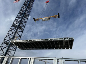 A crane lifts a prefabricated cold-formed steel (CFS) framing section into place, demonstrating the project’s factory-built approach to structural assembly.